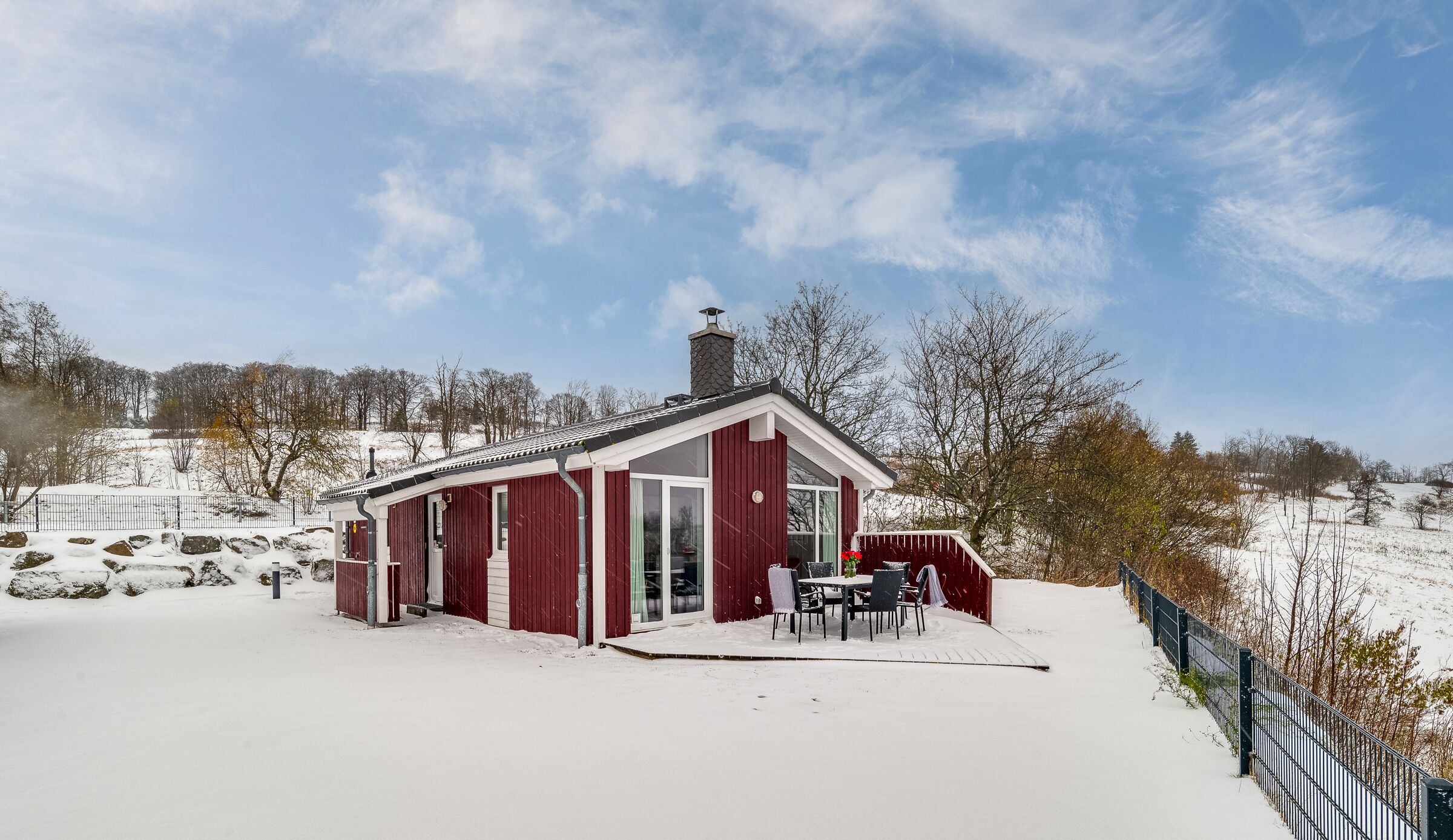 A red wooden cabin with a chimney, a covered patio with outdoor seating, and a snow-covered landscape.