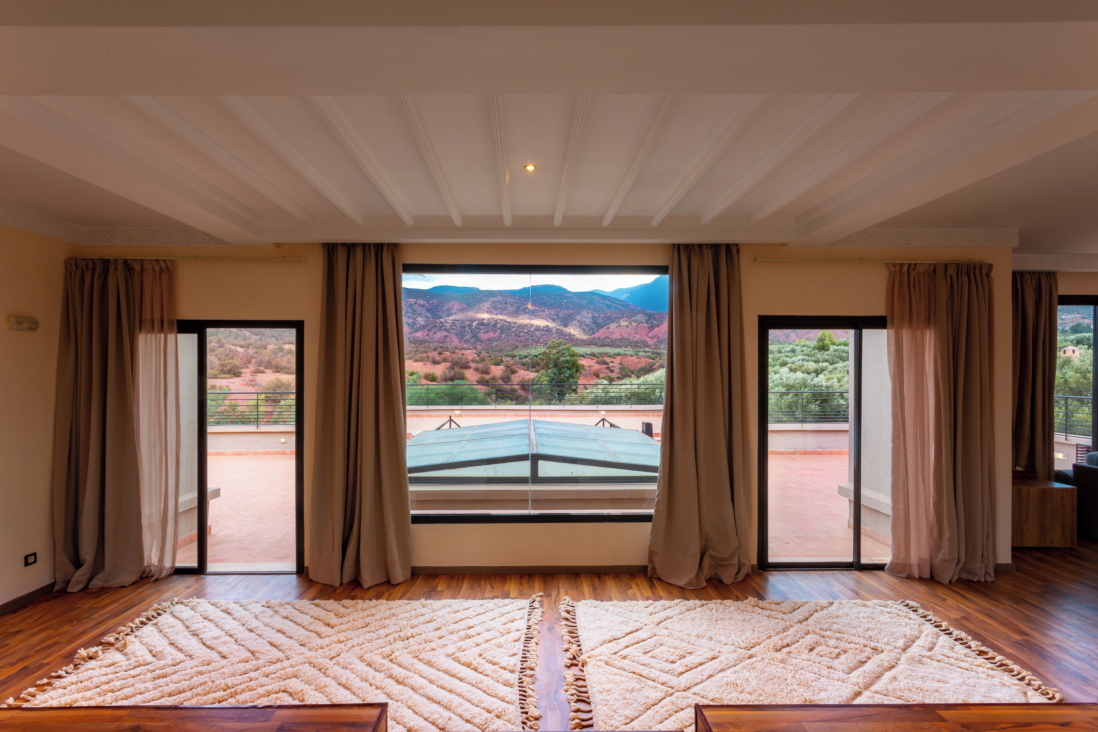 A room with a large window, wooden flooring, and a beige rug.