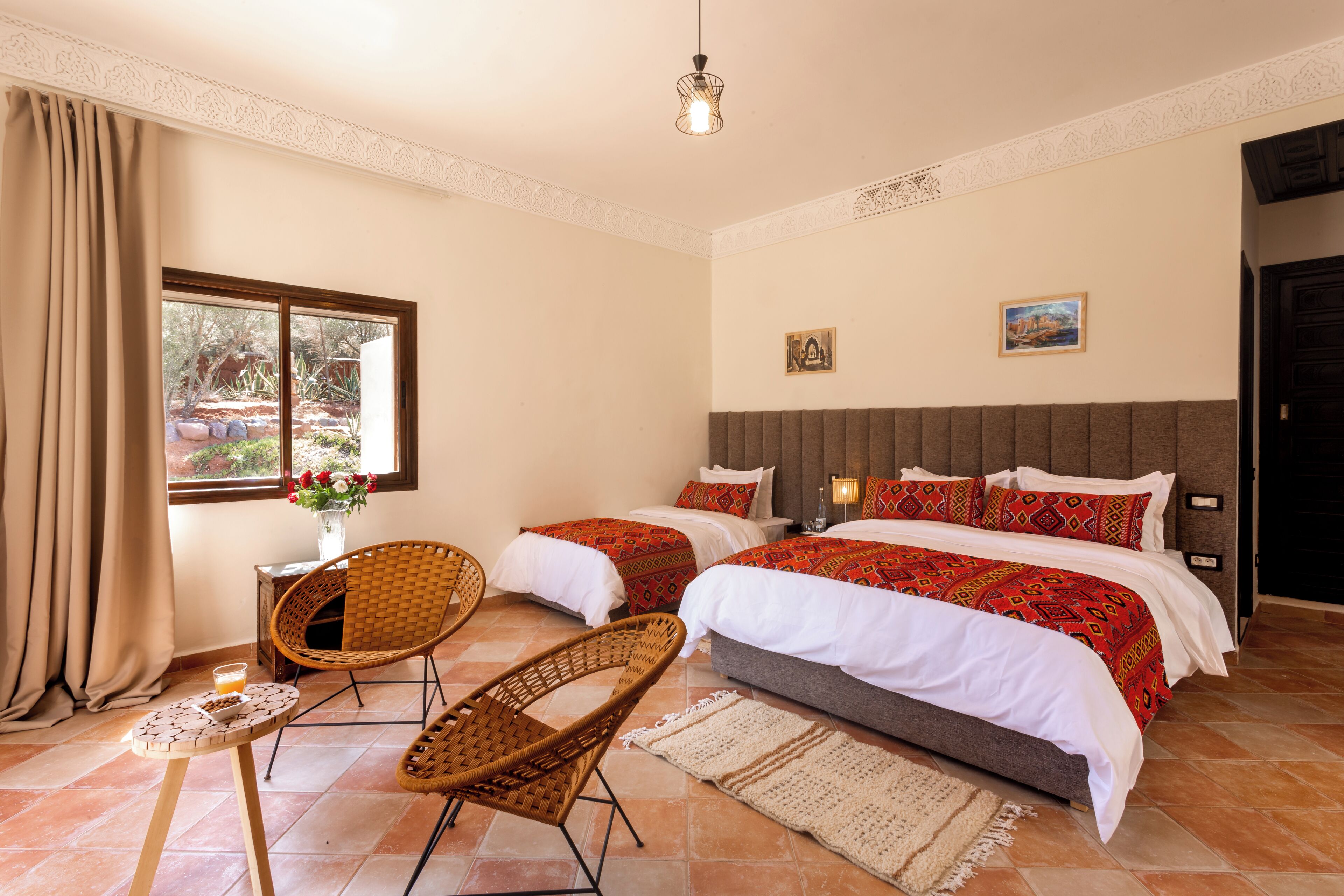 A hotel room with two beds, a sitting area with wicker chairs, a small table with a candle, and a window with curtains.