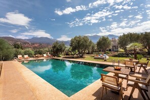 A clear swimming pool with lounge chairs and tables, surrounded by greenery and mountains in the background. - Atlas Luxury Garden (Ouirgane)