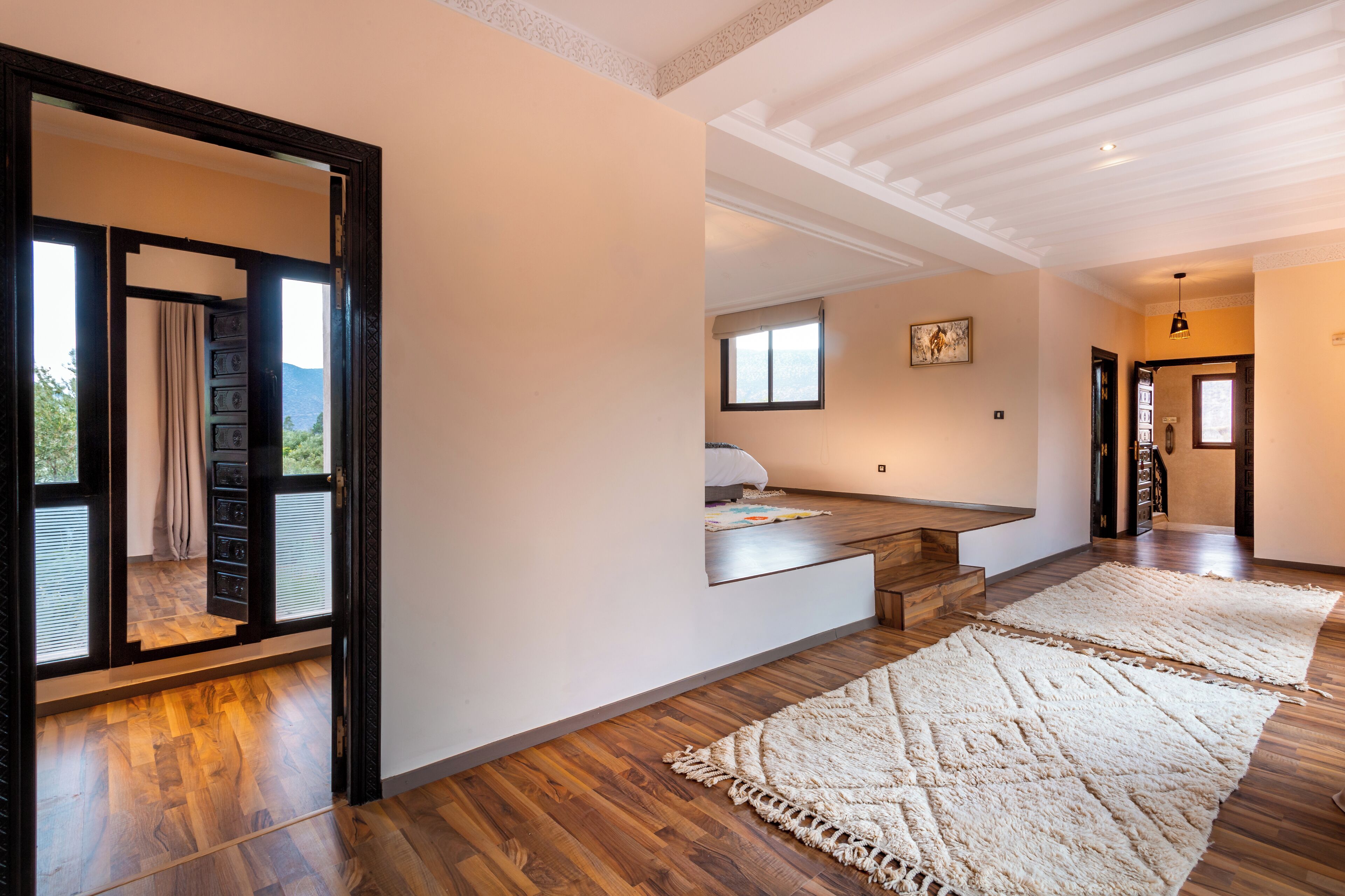 A spacious room with wooden flooring, a large white rug, a bed with a white headboard, and a view of the outdoors through a glass door.