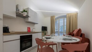 A modern kitchen with white cabinets, a built-in oven, and a coffee machine. There is a dining area with a round table, pink chairs, and a brown sofa. - Ai Portici di Susa - Cozy by Wonderful Italy (Turin)