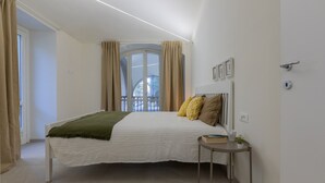 A bedroom with a bed, a bedside table with a book and a plant, a window with curtains, and a door. - Ai Portici di Susa - Cozy by Wonderful Italy (Turin)