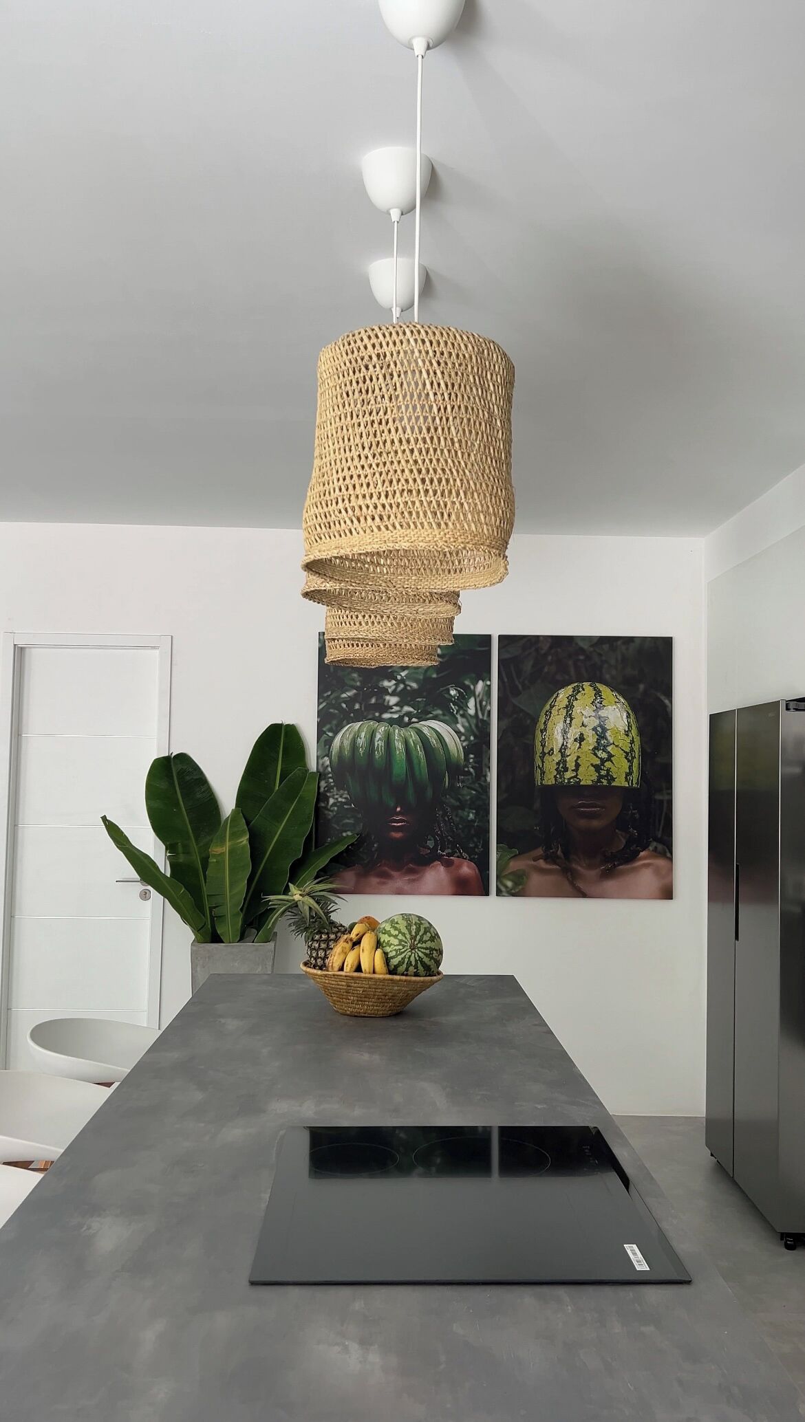 A modern kitchen with a concrete countertop, a woven pendant light, a fruit bowl, and two framed pictures on the wall.