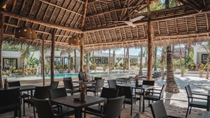 An outdoor dining area with thatched roofs, tables, and chairs, overlooking a pool and lush greenery. - The Oasis Residence (Jambiani)