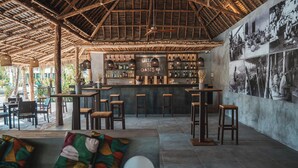 A bar area with wooden stools, a bar counter, and a wall with black and white photographs. - The Oasis Residence (Jambiani)