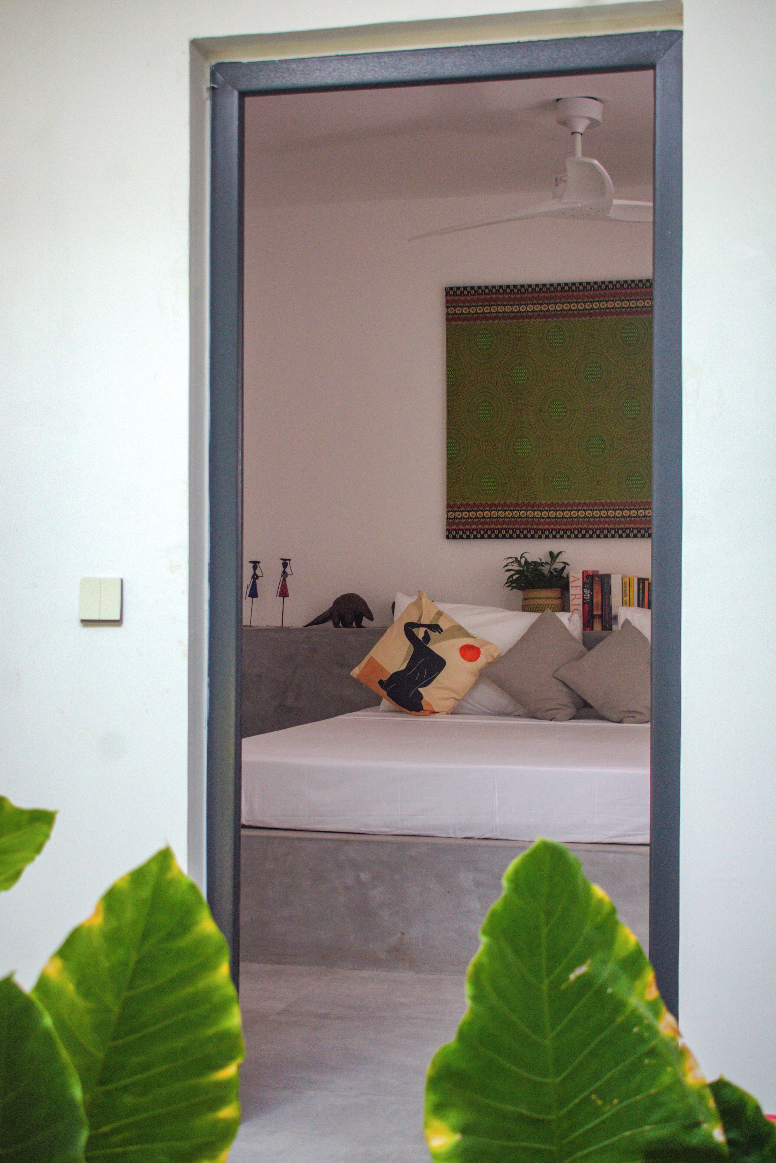 A bedroom with a bed, pillows, a ceiling fan, a wall tapestry, and a small plant.