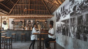 A bar area with wooden stools, a counter with bottles, and a wall with black and white photos. - The Oasis Residence (Jambiani)