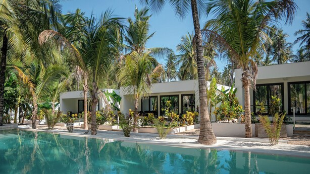 A pool area with palm trees and a building with large windows. - The Oasis Residence (Jambiani)