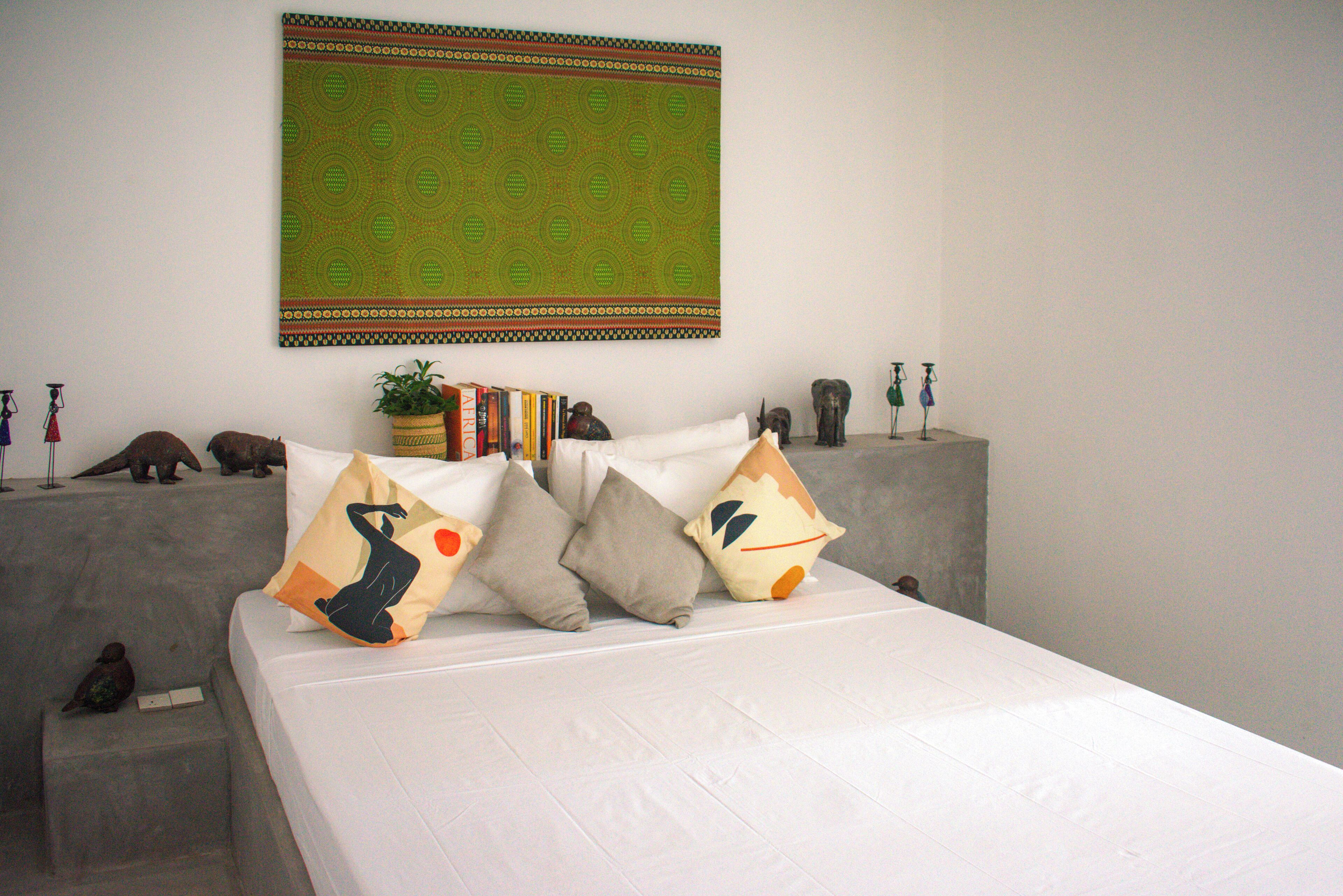 A bed with multiple pillows, a wall hanging, and a small shelf with books and figurines.