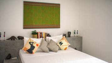 A bed with multiple pillows, a wall hanging, and a small shelf with books and figurines.