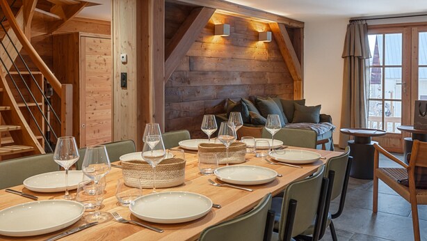 Un coin repas avec une table en bois dressée pour le repas, un escalier et un espace salon confortable avec des coussins.