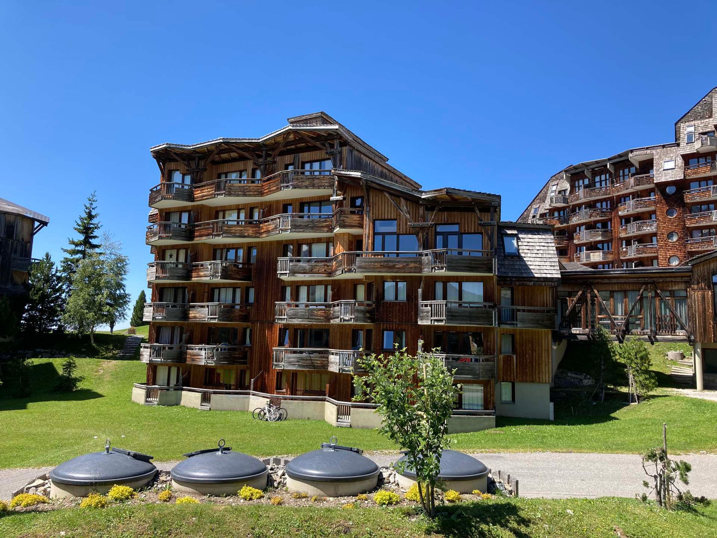 A multi-story wooden building with balconies, a grassy area, and a small tree.