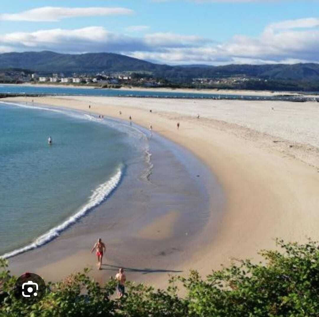 A beach with a curved shoreline, people walking along the beach, and a distant town with mountains in the background.