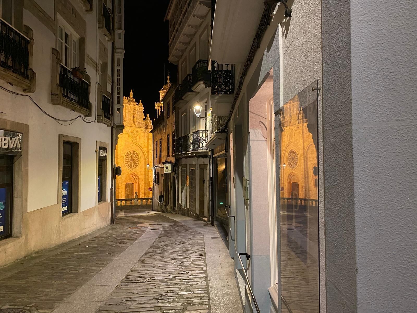 Una calle estrecha flanqueada por edificios, un camino adoquinado y fachadas iluminadas de una iglesia y un edificio con el letrero 'BBVA'.