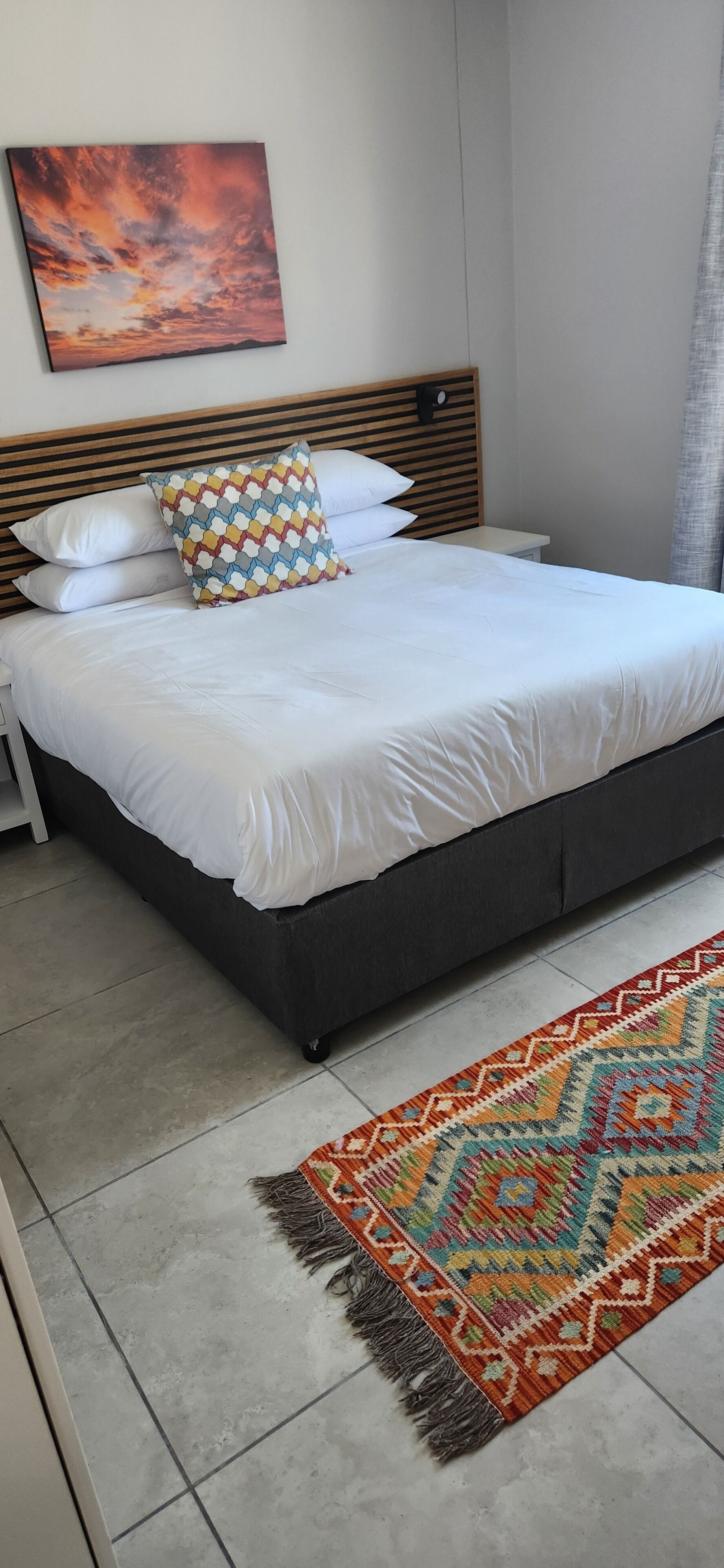 Un lit bien fait, avec du linge de lit blanc, un coussin à motifs et un tapis coloré posé sur un sol carrelé.
