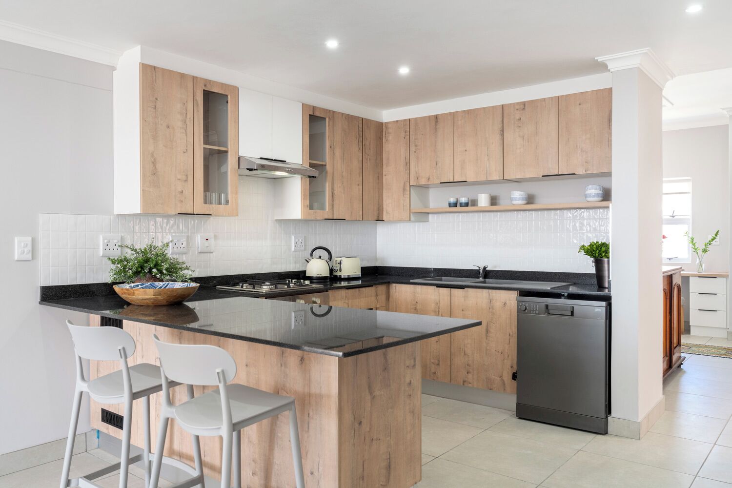 A modern kitchen with wooden cabinets, a central island with bar stools, a built-in dishwasher, and a kettle.