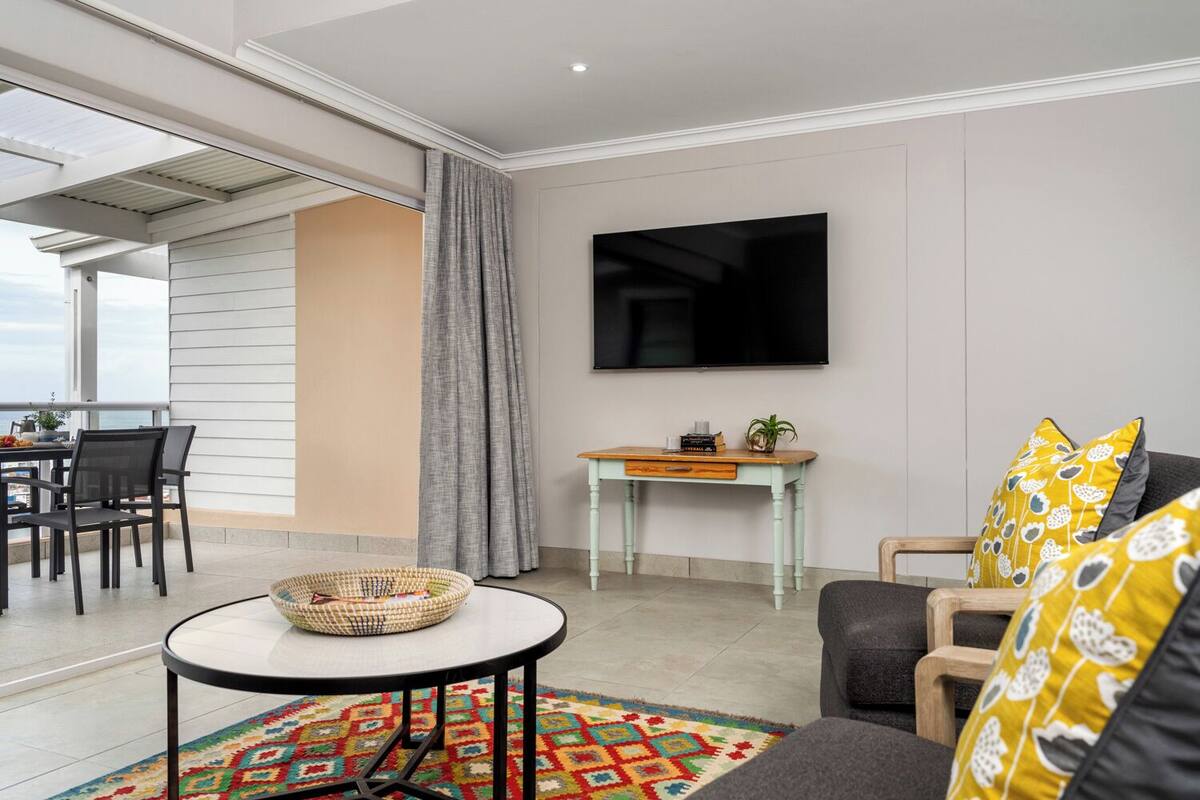 A modern living room with a flat-screen TV, a dining area with chairs, a small table with a plant, and a colorful rug.