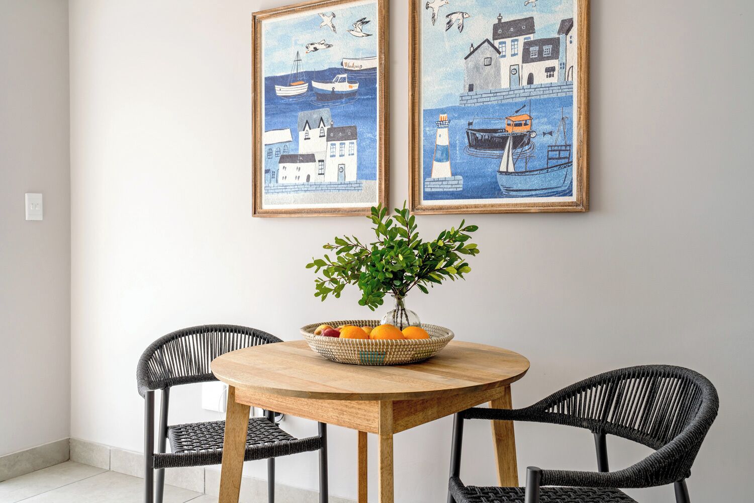 A dining area with a round wooden table, two black wicker chairs, and a vase with green foliage and fruit. Two framed artworks depicting coastal scenes are hung on the wall.