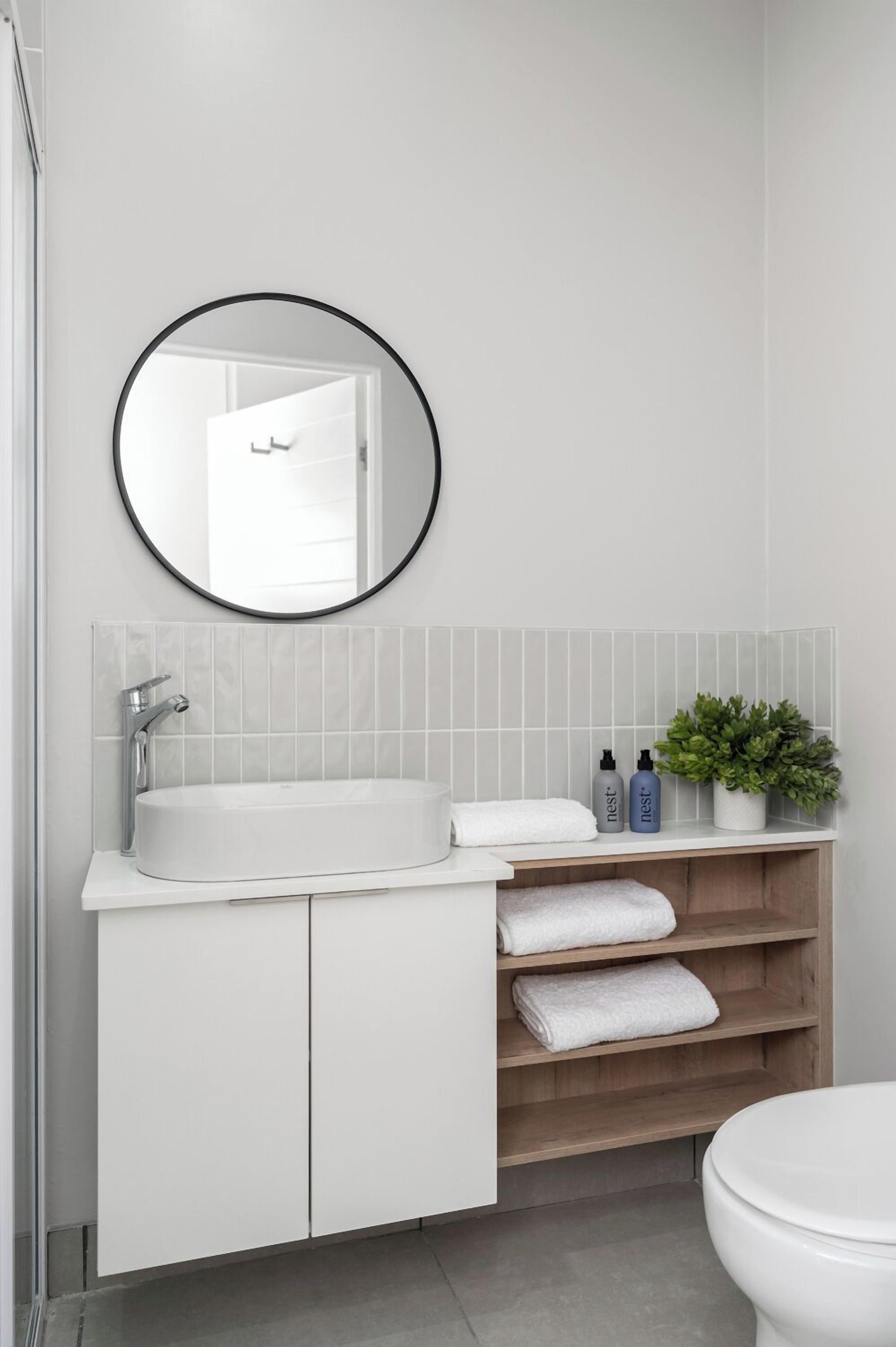A bathroom with a white sink, a round mirror, and a wooden shelf with towels.