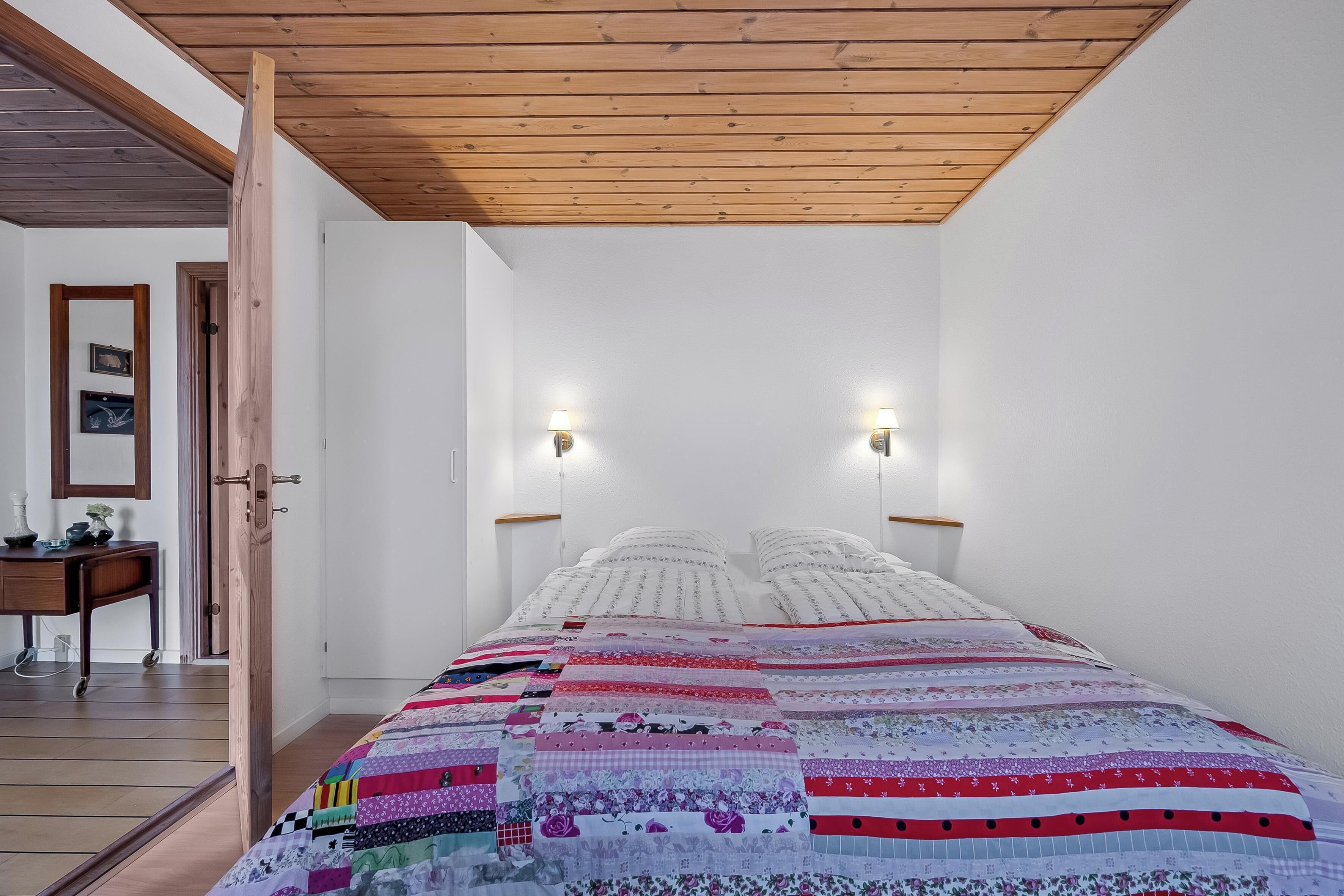 A bedroom with a wooden ceiling, white walls, a quilted bedspread, two bedside lamps, and a wooden nightstand with a lamp.