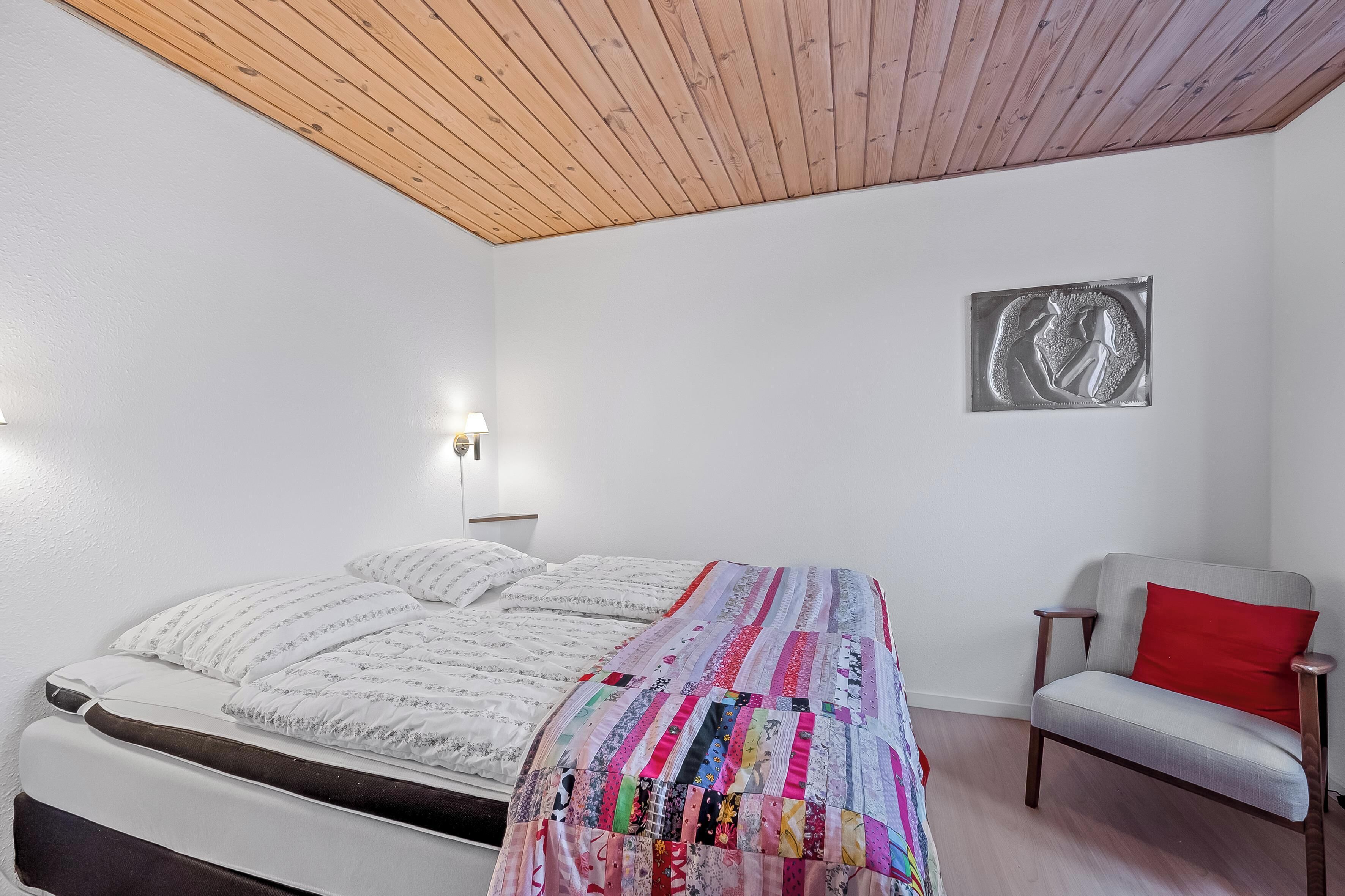 A bedroom with a wooden ceiling, a bed with a colorful quilt, a gray chair with a red pillow, and a wall-mounted light fixture.