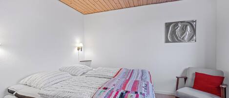 A bedroom with a wooden ceiling, a bed with a colorful quilt, a gray chair with a red pillow, and a wall-mounted light fixture.