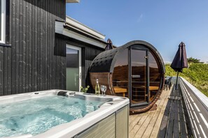 A hot tub outside a wooden structure with a deck and a glass-enclosed seating area.