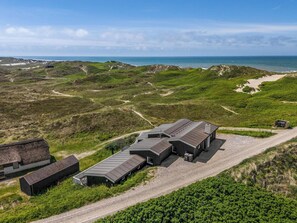 A modern building with a gravel driveway, situated on a grassy hill with a view of the sea and wind turbines in the distance.