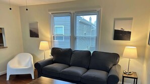 A living room with a grey sofa, a white chair, a wooden coffee table, two floor lamps, and a window with blinds.