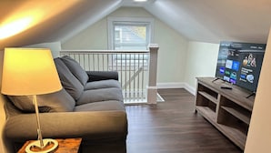 A cozy living room with a grey sofa, a small wooden table, a lamp, and a TV displaying streaming services.