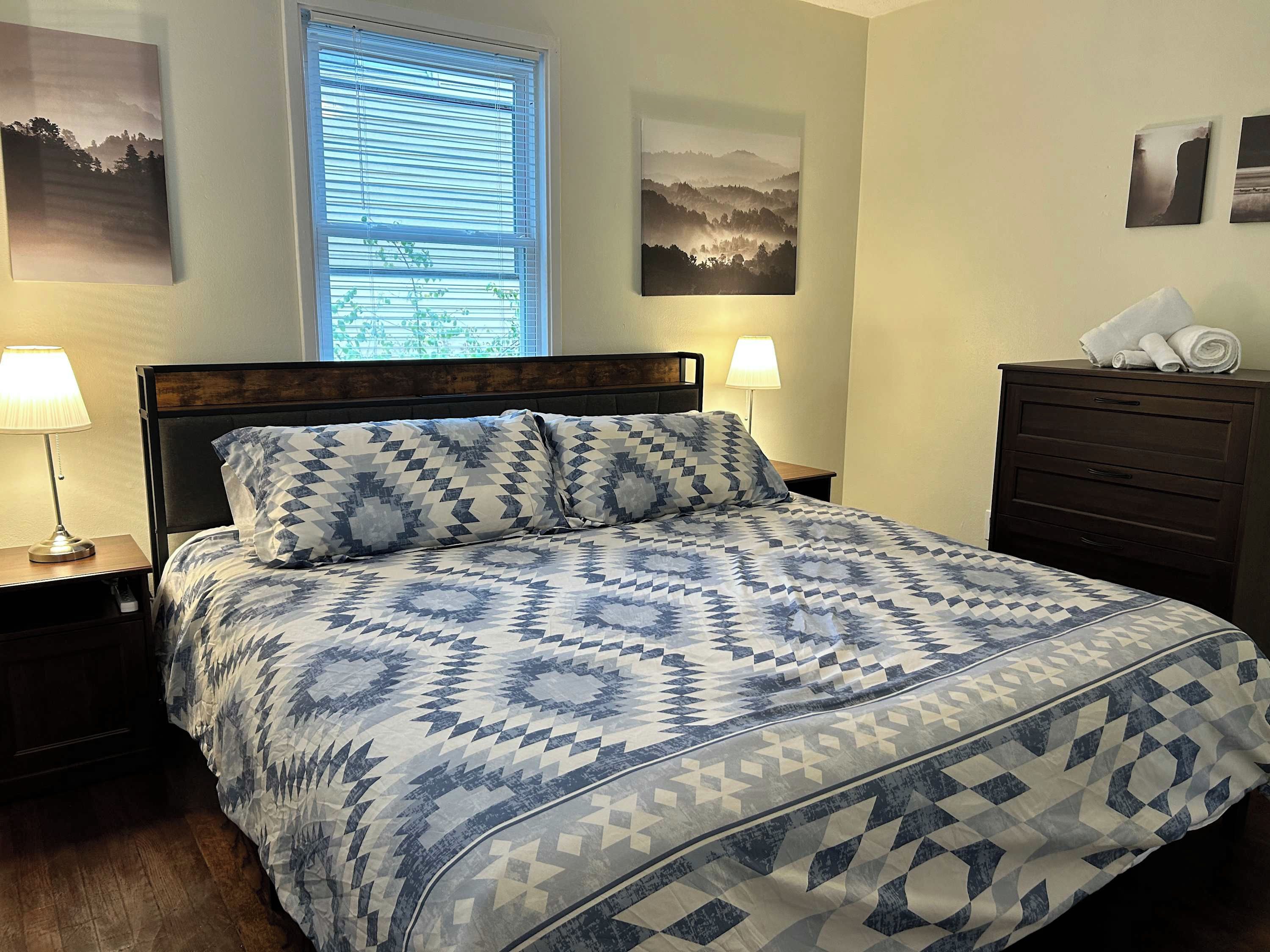 Una cama bien hecha con una colcha de diseño geométrico, cabecera de madera y mesita de noche con lámpara. Se ve una ventana con persianas, un aparador con toallas y dos cuadros enmarcados en la pared.