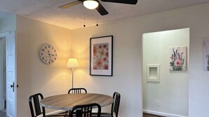A dining area with a round table, four chairs, a wall clock, a floor lamp, and two framed artworks.