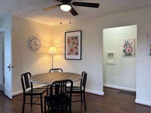 A dining area with a round table, four chairs, a wall clock, a floor lamp, and two framed artworks.
