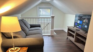 A cozy living room with a grey sofa, a small wooden table, a lamp, and a TV displaying streaming services.