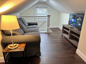 A cozy living room with a grey sofa, a small wooden table, a lamp, and a TV displaying streaming services.