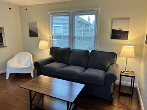 A living room with a grey sofa, a white chair, a wooden coffee table, two floor lamps, and a window with blinds.
