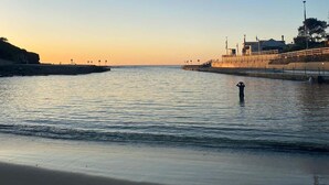 Una playa con aguas tranquilas, una persona de pie en el agua y un edificio con una bandera a la derecha.