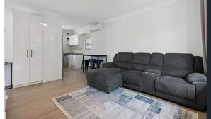 Un salon moderne avec un canapé d’angle gris, un petit pouf et un tapis à motifs. On aperçoit à l’arrière-plan un coin cuisine avec des meubles blancs et une table à manger.