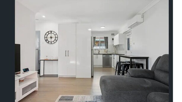 Un salon moderne avec un canapé gris, un téléviseur à écran plat et un coin cuisine à l’arrière-plan.