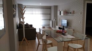 A modern living room with a glass dining table, white chairs, a brown sofa, and a wooden coat rack. - APARTMENTS NAJERA. Comfort for an exceptional rest. (Nájera)