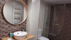 A modern bathroom with a round mirror, a white sink, a toilet, and a shower area. - APARTMENTS NAJERA. Comfort for an exceptional rest. (Nájera)