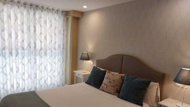 A neatly made bed with a headboard, two bedside tables with lamps, and a window with patterned curtains. - APARTMENTS NAJERA. Comfort for an exceptional rest. (Nájera)
