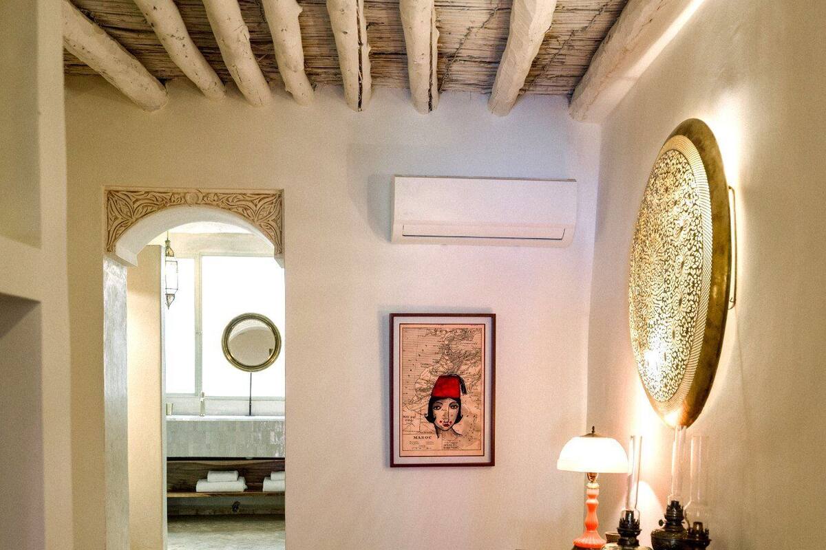 A hallway with a wooden ceiling, a green armchair, a patterned rug, and a table with a lamp and decorative items.