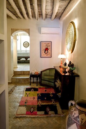 Un couloir avec un plafond en bois, un fauteuil vert, un tapis à motifs et une table sur laquelle se trouvent une lampe et des objets décoratifs.