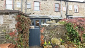 A stone building with a small garden area, a wooden door, and a window with a shutter. - Three Bed Cottage in the heart of the Peaks (Edale)