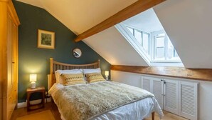 A bedroom with a sloped ceiling, a bed with white bedding, a nightstand with a lamp, and a window with shutters. - Three Bed Cottage in the heart of the Peaks (Edale)