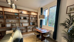 Un salon chaleureux avec une bibliothèque en bois, une table à manger entourée de chaises, un canapé et une grande fenêtre agrémentée de rideaux.
