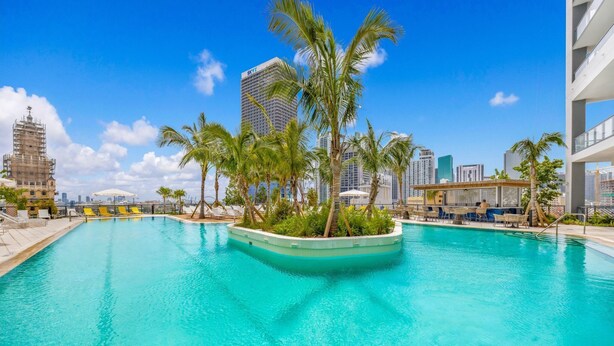 Una piscina rodeada de palmeras, edificios modernos y un cielo azul despejado.