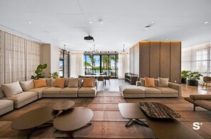 A spacious living area with a large sectional sofa, a coffee table, and a dining table. There are potted plants and a view of palm trees through large windows.